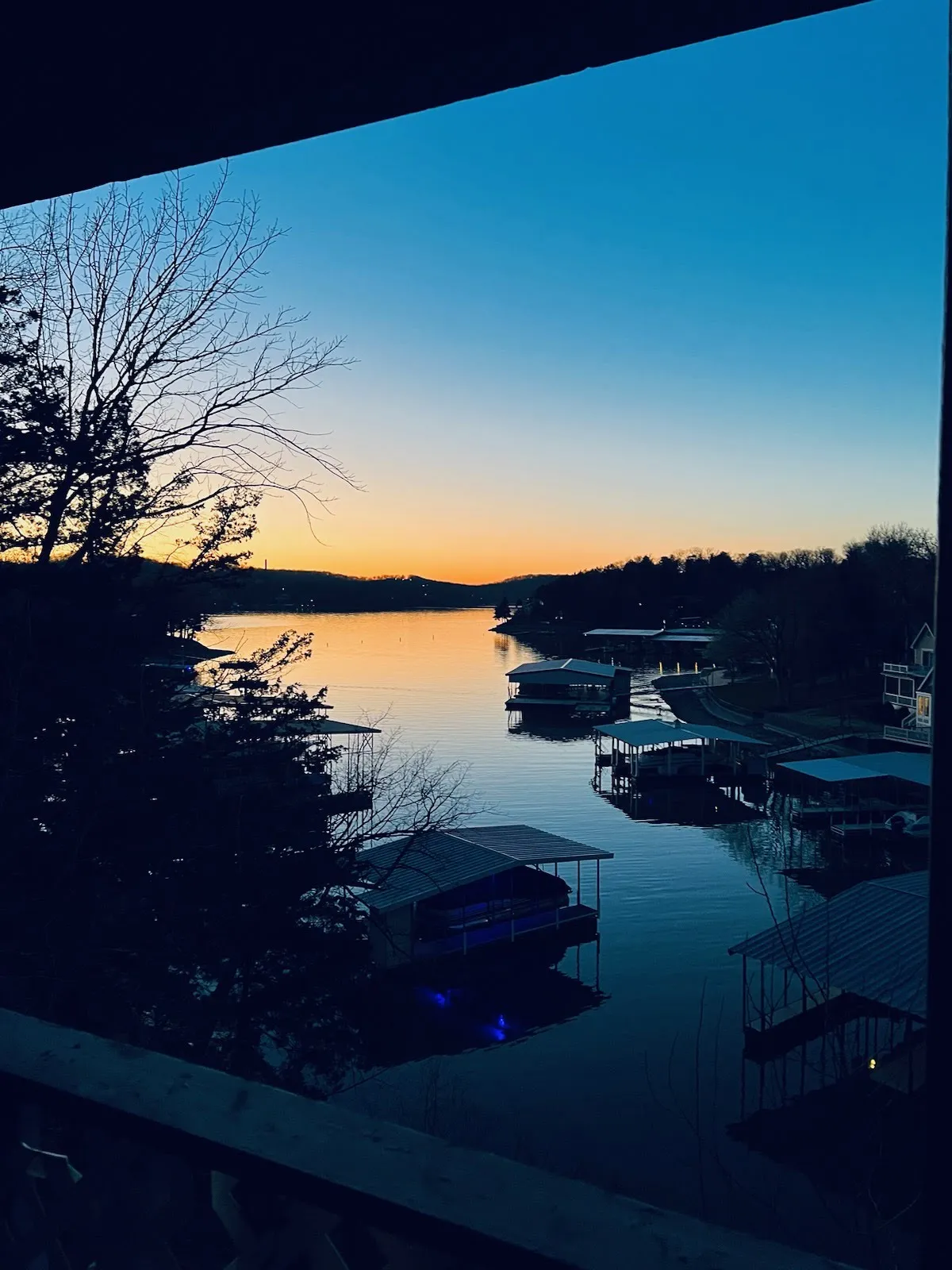 A serene view of the calm lake surface, boathouses along the shore, and silhouetted trees all set against vibrant sunset colors. The scene conveys peaceful waterfront relaxation, with activities like boating, fishing, and simply enjoying the natural beauty at dusk.