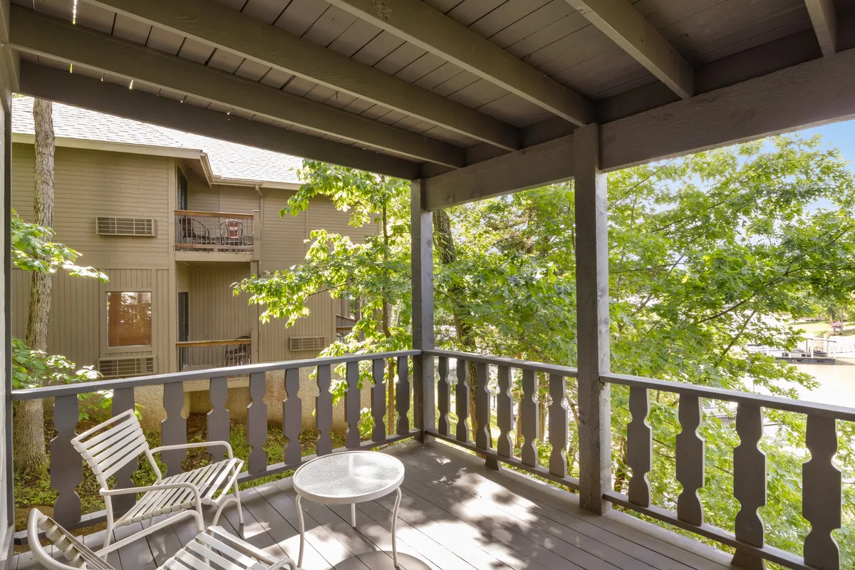 A spacious balcony with a wooden railing overlooks the lake, with an outdoor seating set and lush green trees. The view emphasizes the connection between the home and its natural surroundings, ideal for sunrise coffees or evening gatherings.