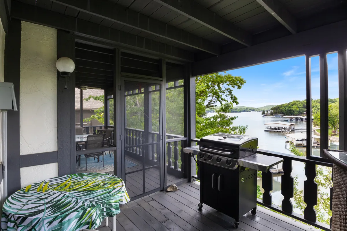 An outdoor porch scene featuring a gas grill, a table with a tropical tablecloth, and a serene view of the lake. The setup is relaxed yet expertly arranged for summer barbecues and alfresco dining.