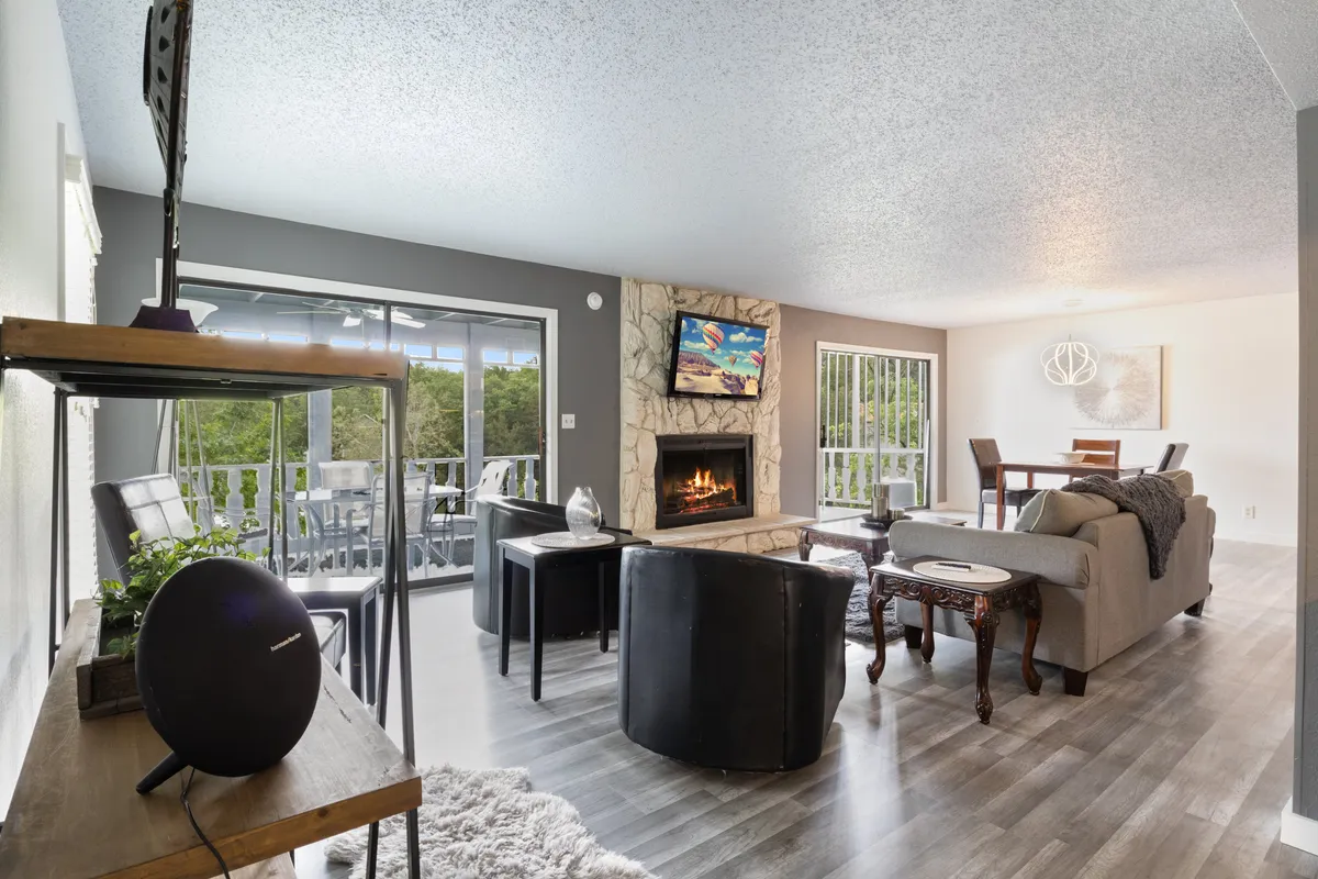 A spacious living area with a prominent fireplace, a large flat screen TV mounted above, plush seating arranged for conversation and relaxation, and expansive windows revealing beautiful lake views. The adjacent dining table suggests fluid movement between lounging and dining, reinforcing the open-plan layout.
