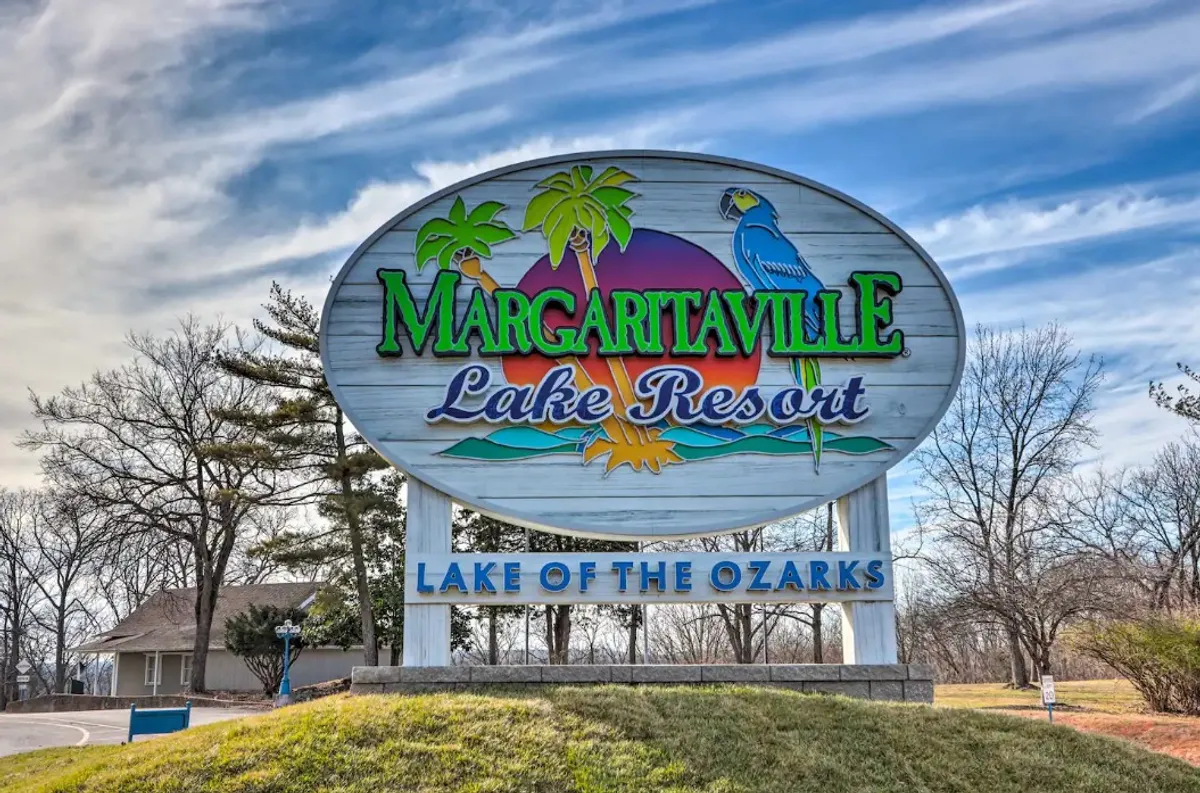 A vibrant resort entrance scene with a festive sign, palm trees, and colorful parrots. The atmosphere is lively, hinting at the community's spirited vibe and proximity to lakeside activities and events.
