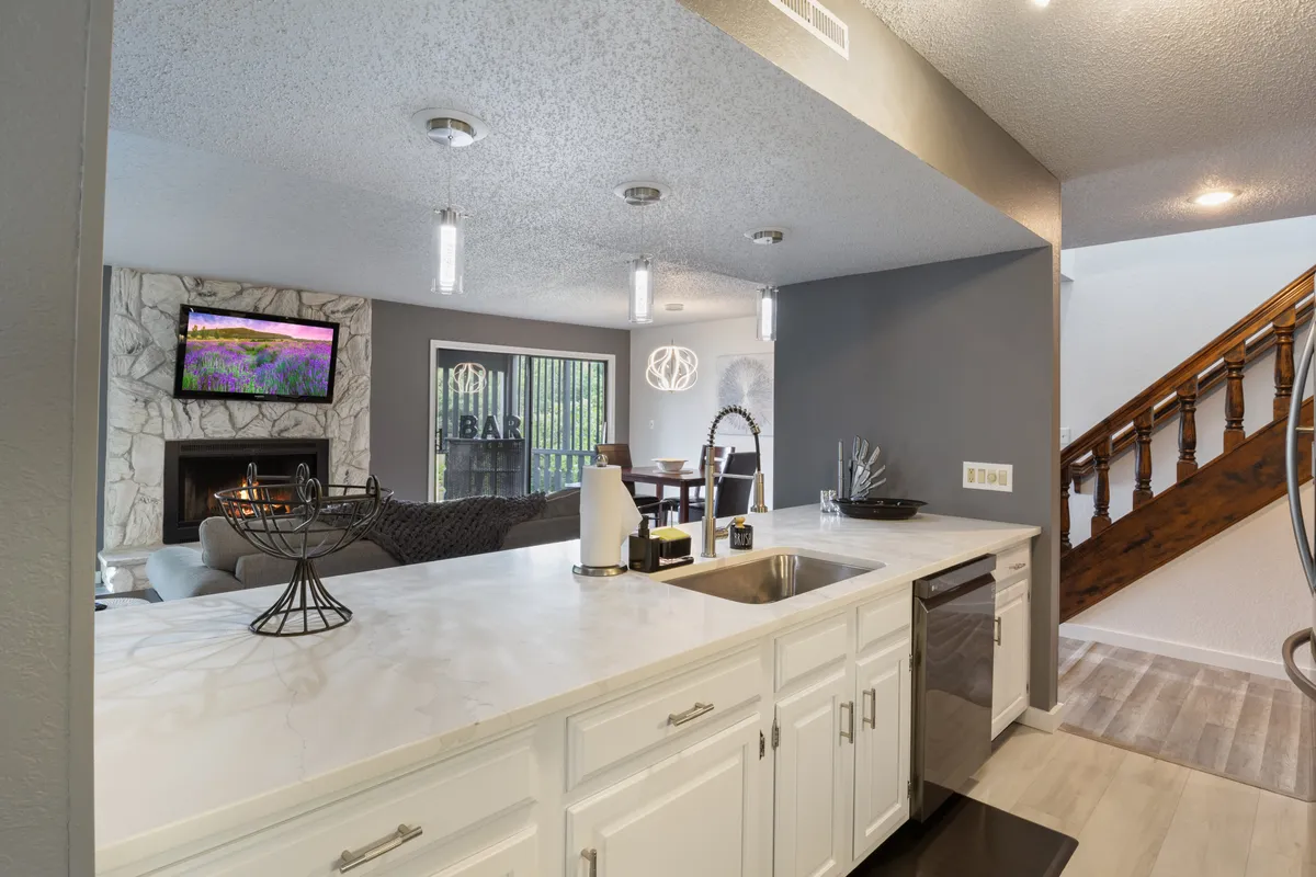 A welcoming open living space that seamlessly connects the kitchen and living area. The arrangement includes a kitchen island, a comfortable seating area, a fireplace, and a flat screen TV, evoking a sense of easy, sociable flow.
