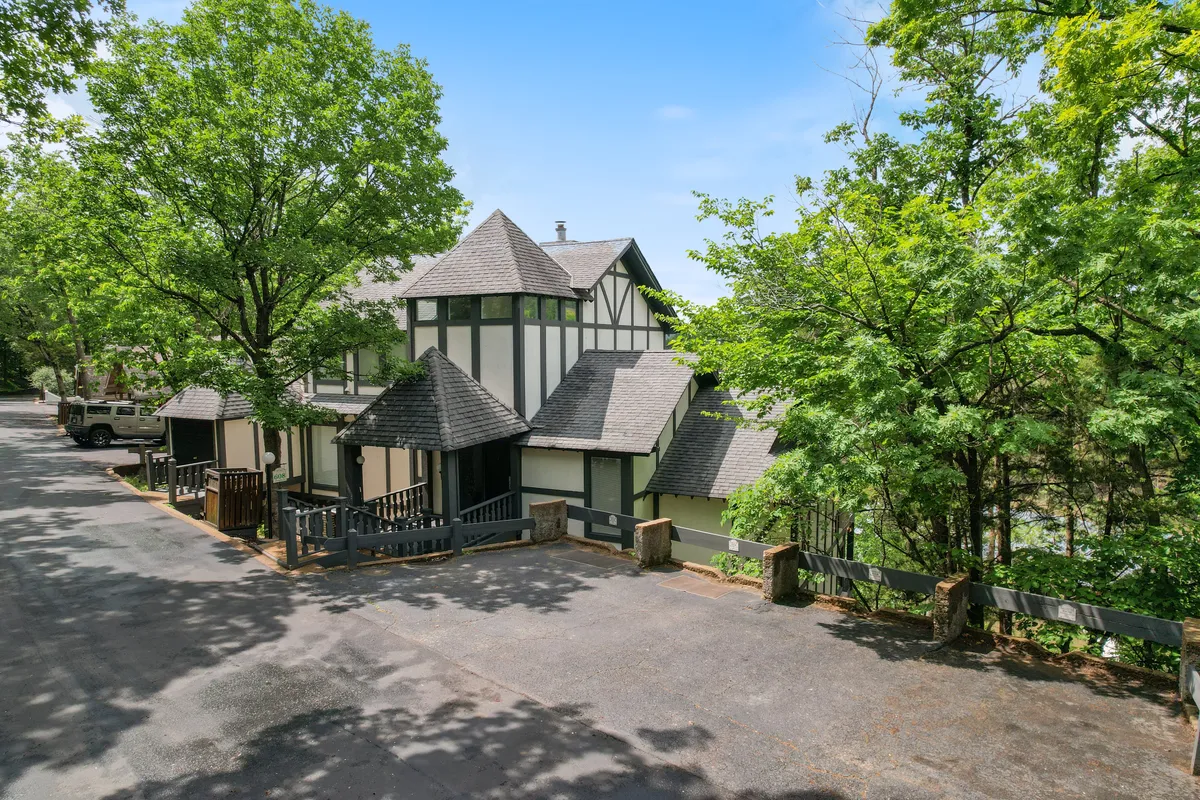 A welcoming exterior view of the Swiss House, showcasing its distinctive Tudor-style architecture surrounded by lush greenery. The driveway leads up to the home, hinting at its private, exclusive lakeside setting. The porch and open surroundings evoke a sense of relaxation and connection with nature.