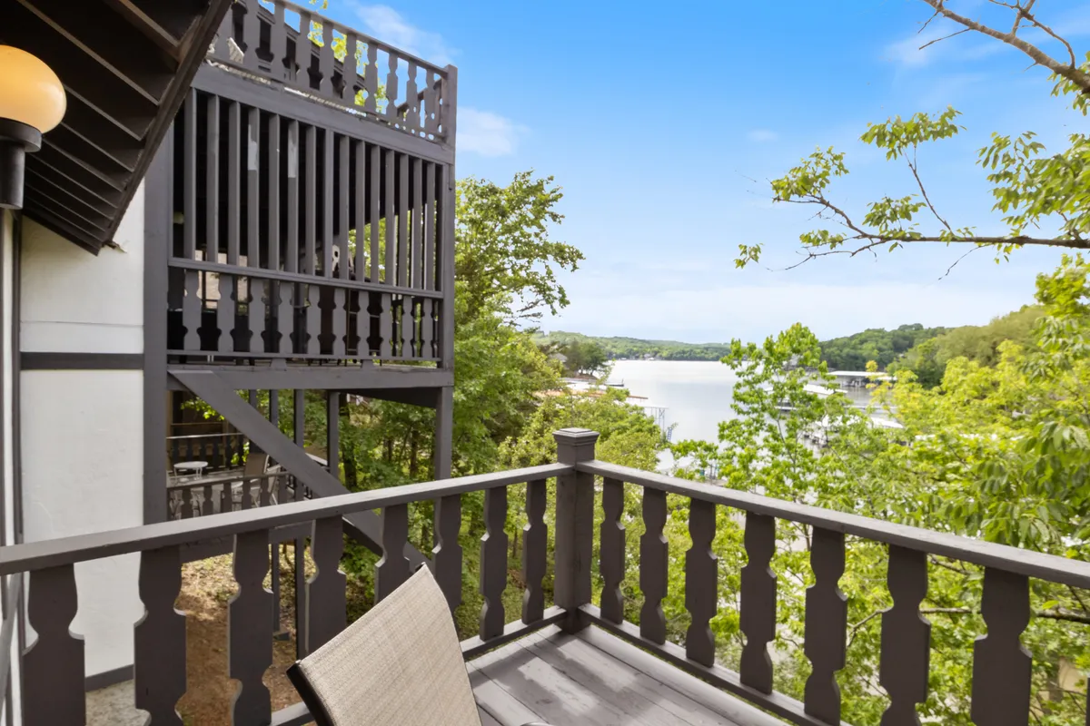 A peaceful exterior view featuring a wooden balcony railing overlooking the water, surrounded by lush greenery. The setting invites guests to relax, socialize, and take in the tranquil lake scenery.