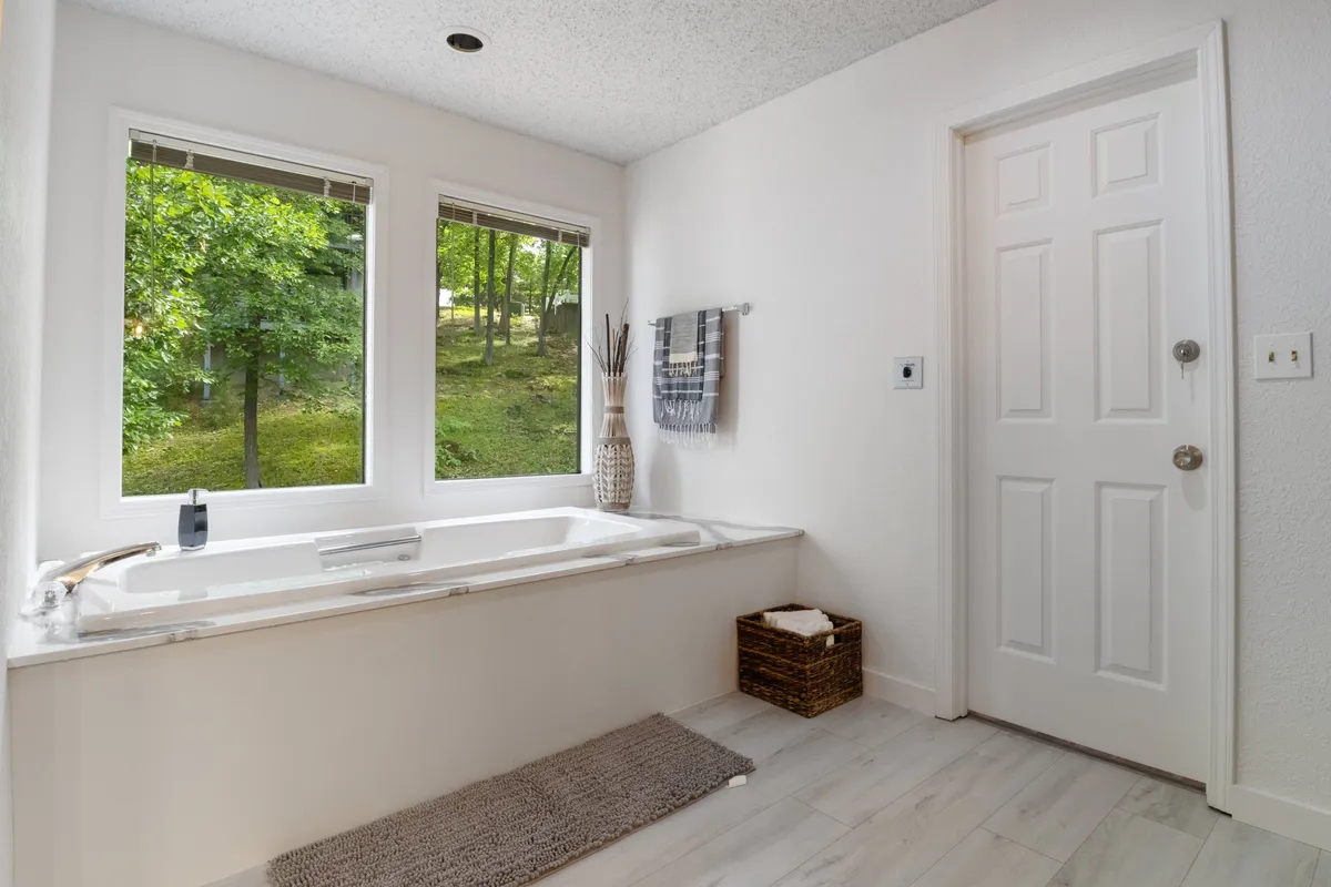 A spa-like bathroom featuring a luxurious bathtub positioned by large windows, allowing natural light to filter in. The space is accented with a decorative vase, towel rack, and calming, modern finishes, creating an inviting atmosphere for relaxation.